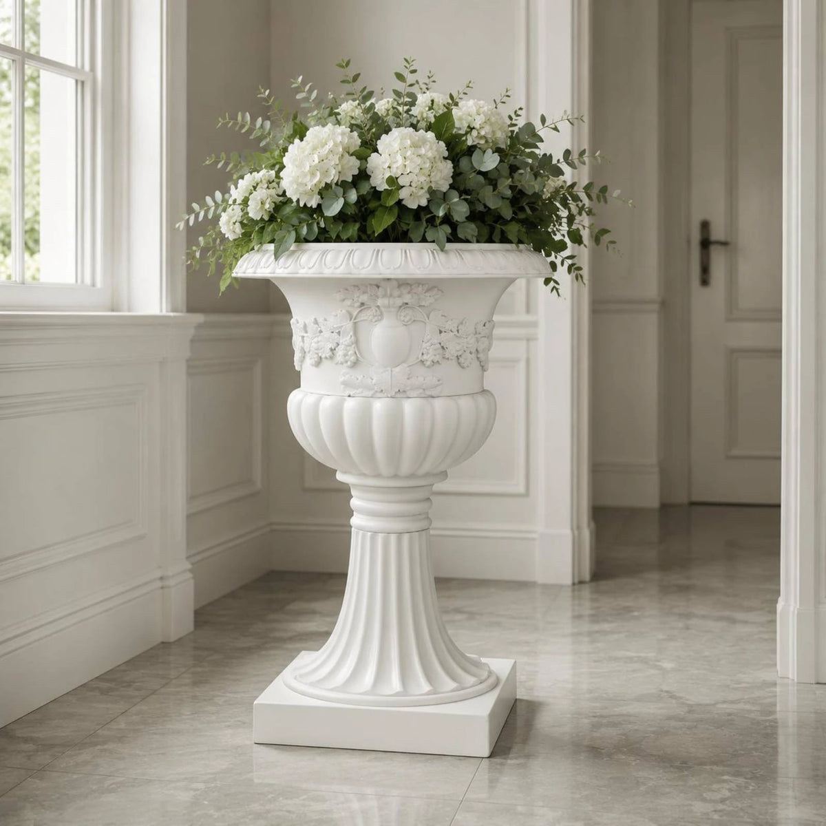 Maceta blanca de relieve floral con pedestal y flores blancas.
