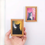 Mano sosteniendo un imán con retrato de gato negro sobre fondo ocre.