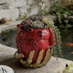 Maceta roja con forma de corazón anatómico sostenido por una mano dorada, con suculentas.