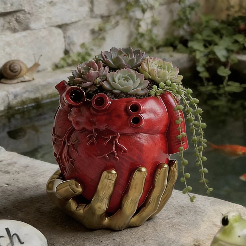 Maceta roja con forma de corazón anatómico sostenido por una mano dorada, con suculentas.