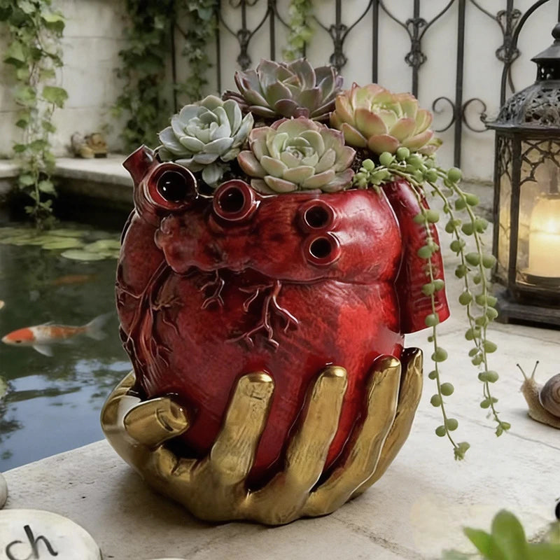 Maceta de corazón rojo y mano dorada en un jardín, con suculentas.